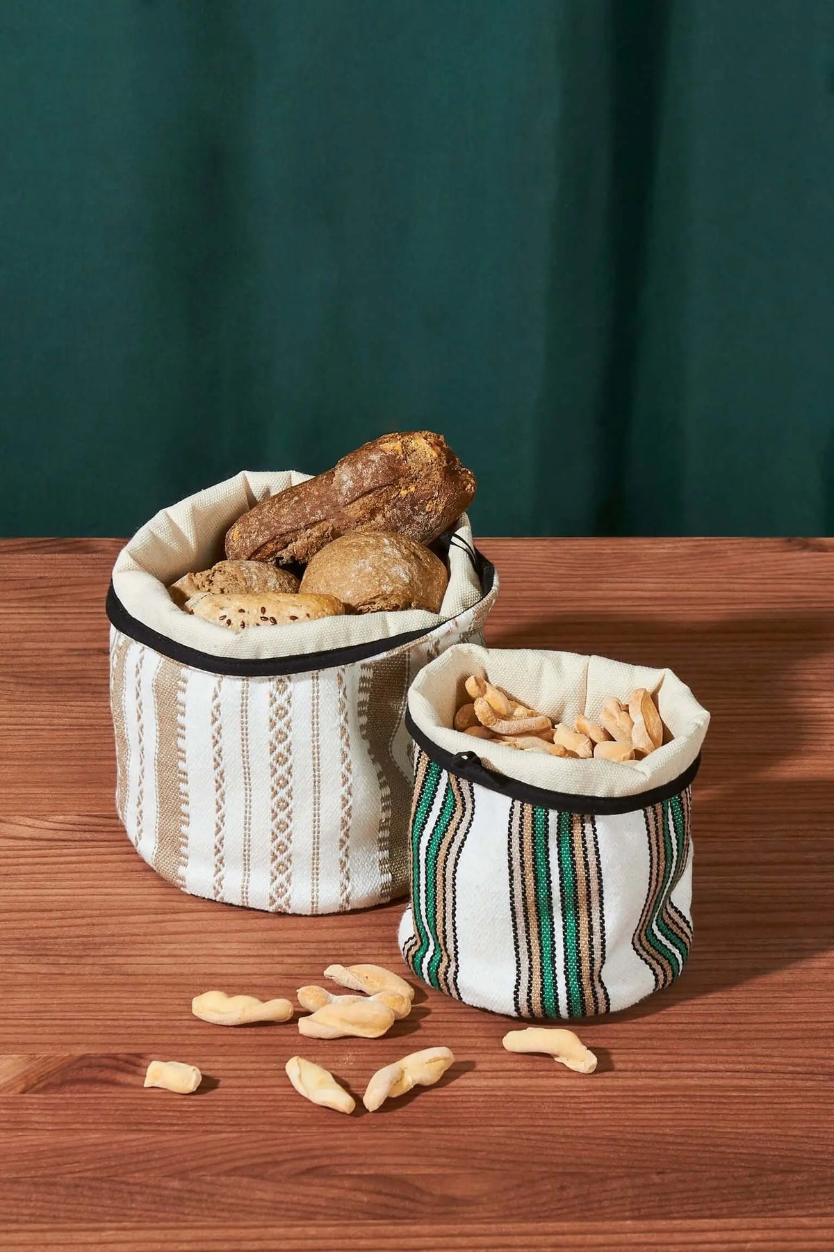 Dos cestos de tela con rayas verdes y beige, con bordes negros, llenos de pan y snacks sobre una mesa de madera.