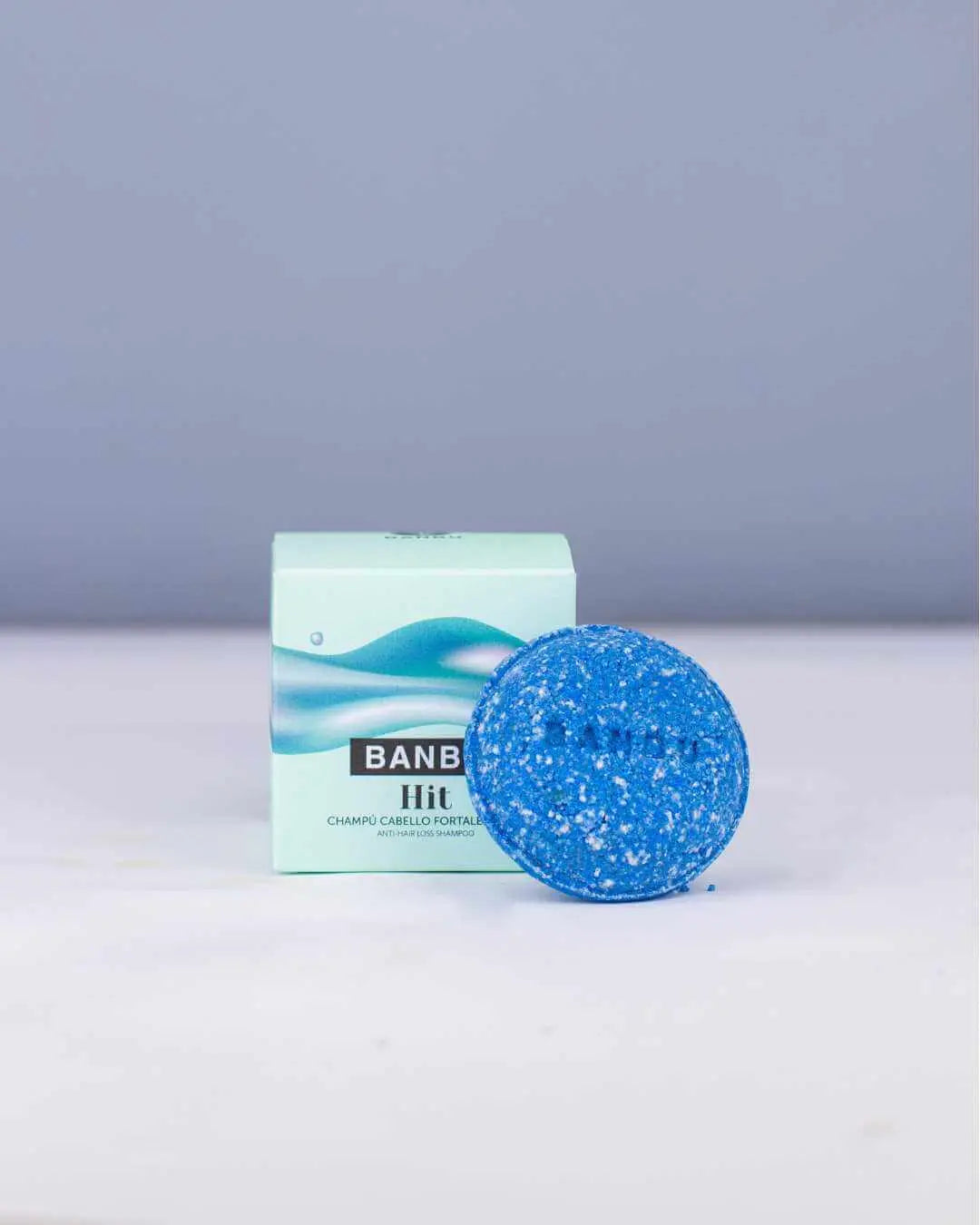 Pastilla de champú sólido azul y caja de cartón verde claro de la marca Banbu sobre superficie blanca.