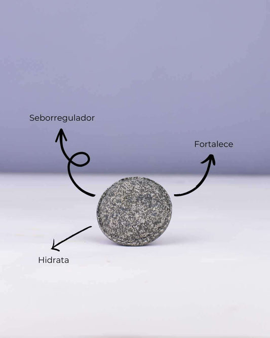 Champú sólido gris oscuro Mare de Banbu sobre fondo claro, con flechas indicando sus beneficios: regula, hidrata y fortalece.