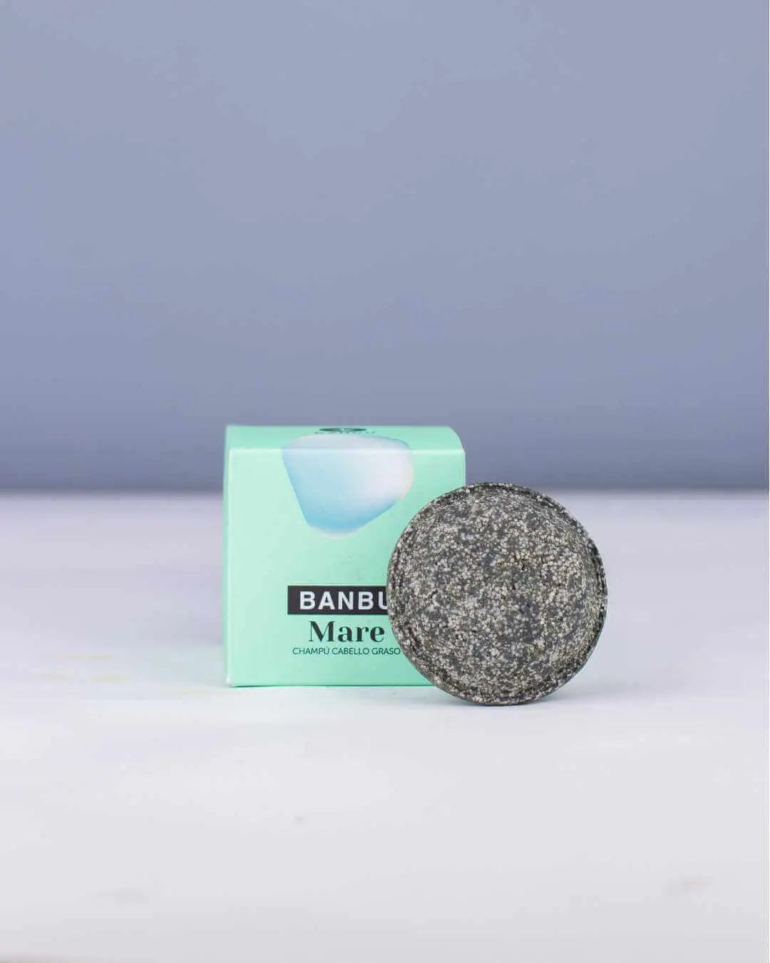 Champú sólido gris texturizado frente a caja verde menta de marca Banbu, sobre superficie blanca y fondo azul suave.
