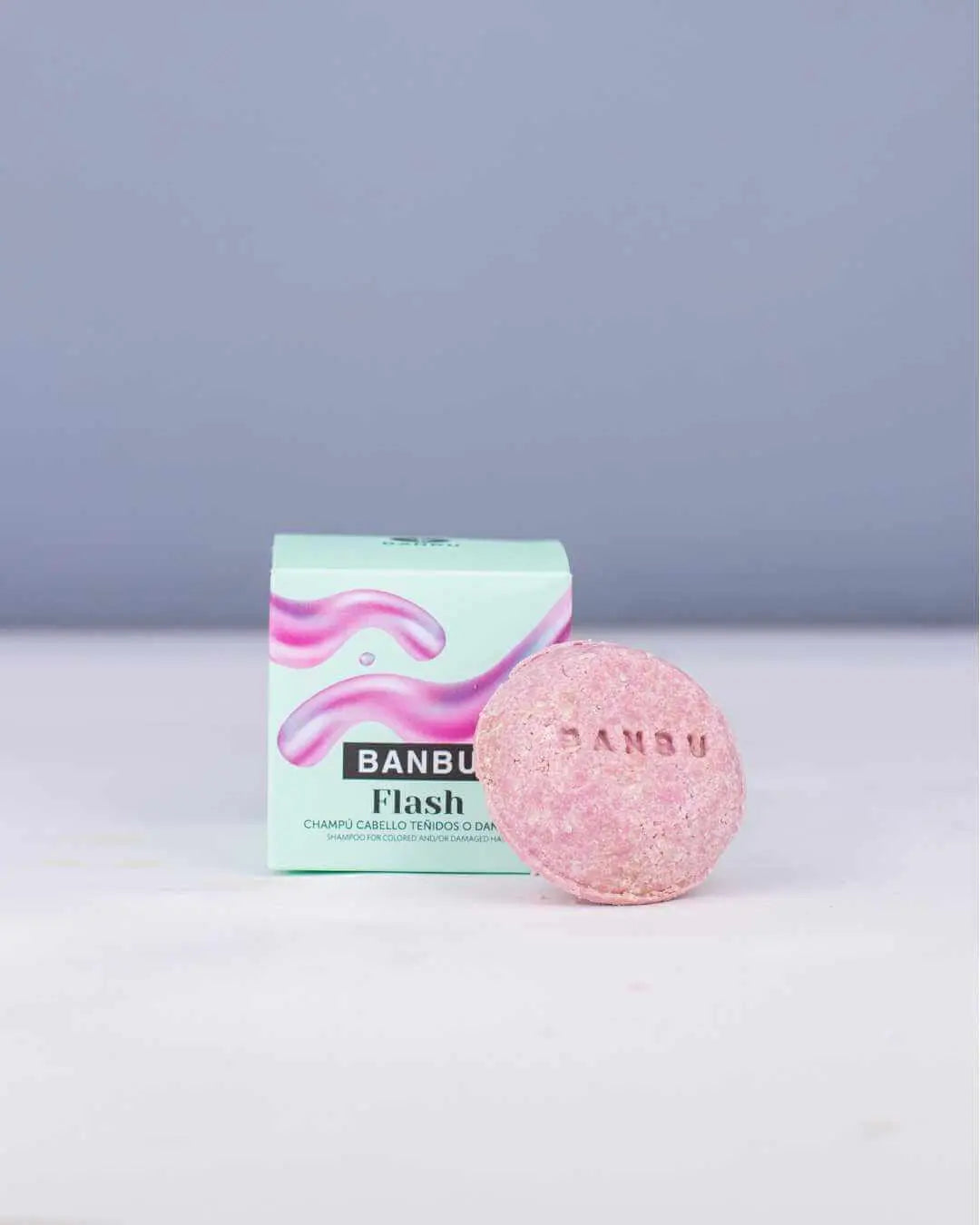 Champú sólido rosa con logo Banbu y caja verde menta sobre fondo claro. Producto ecológico para cabello dañado.