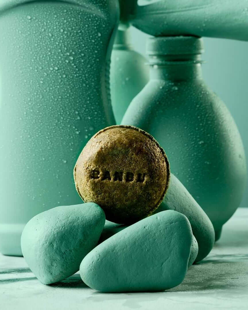 Champú sólido redondo Banbu sobre piedras turquesas, frente a botellas de plástico con gotas de agua. Fondo minimalista.
