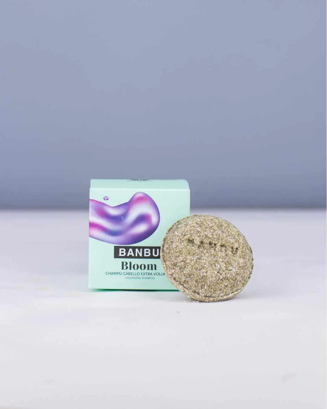 Champú sólido Banbu Bloom de textura granulada frente a su caja de cartón verde claro con detalle morado iridiscente.
