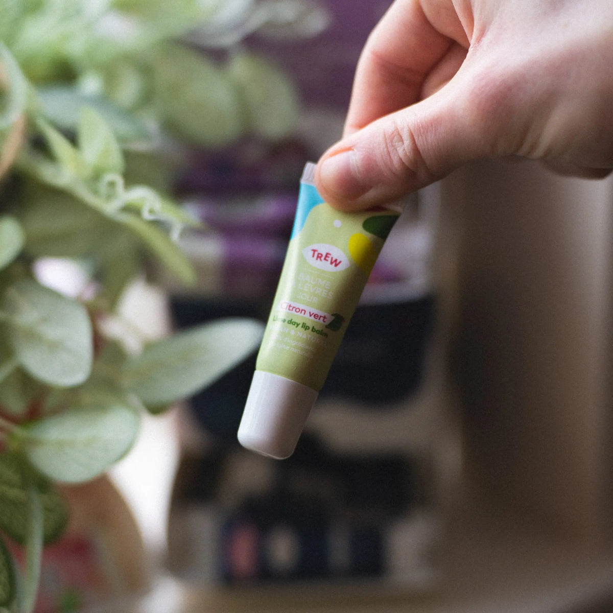 A hand holds a lime green tube of Trew Baume Jour Citron Vert lip balm against a blurred botanical background.