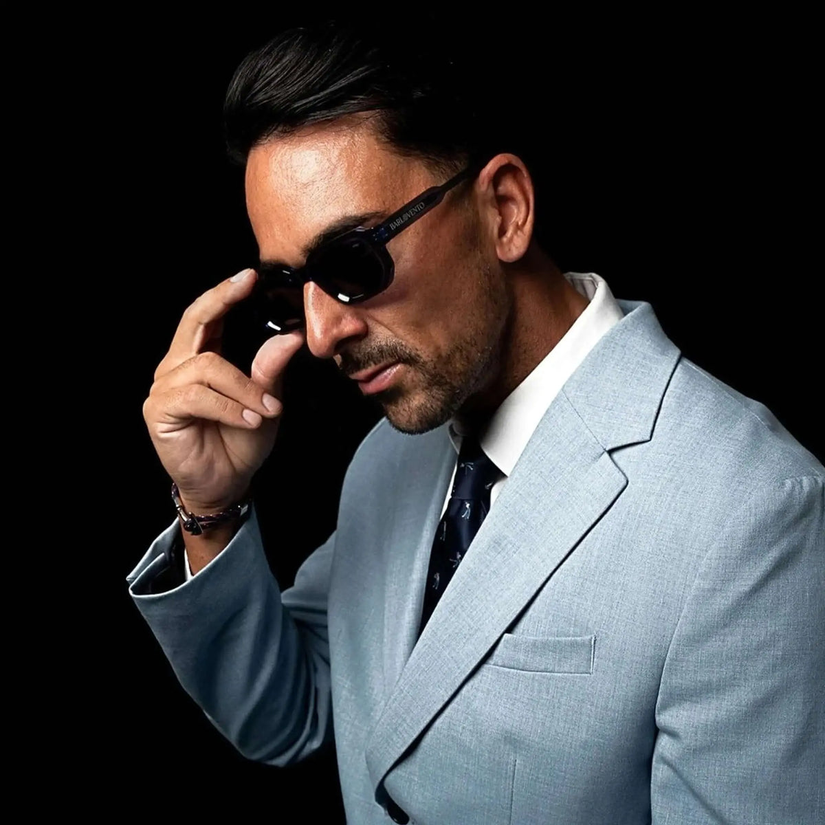 Hombre con americana azul claro y gafas de sol de montura azul intenso y lentes oscuras, sobre fondo negro.