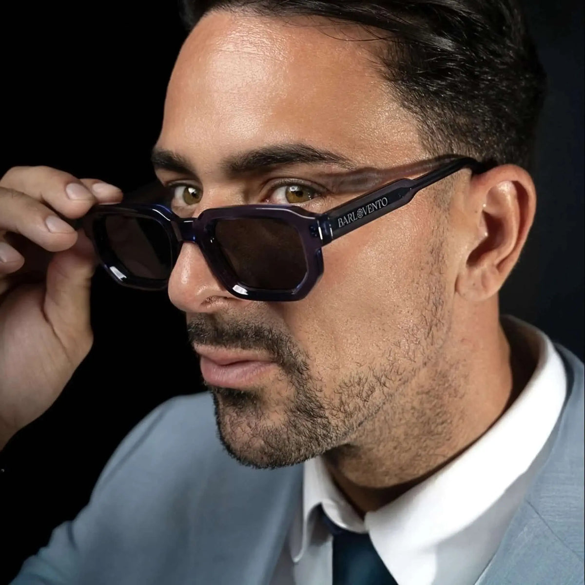 Gafas de sol de montura cuadrada en azul traslúcido con lentes oscuras, puestas por un hombre con traje.