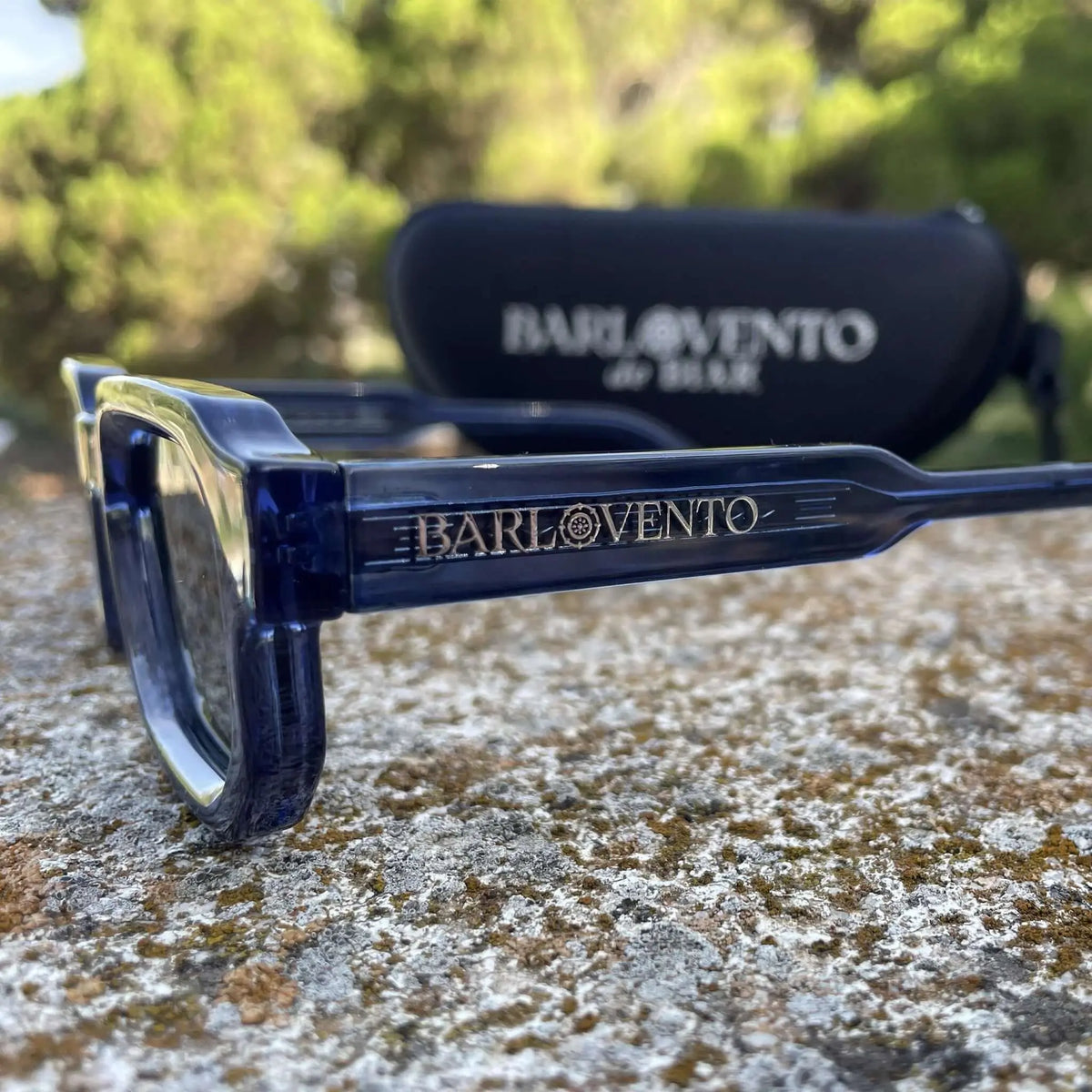 Gafas de sol de montura rectangular en acetato azul traslúcido y lentes oscuras, apoyadas sobre una superficie rugosa.