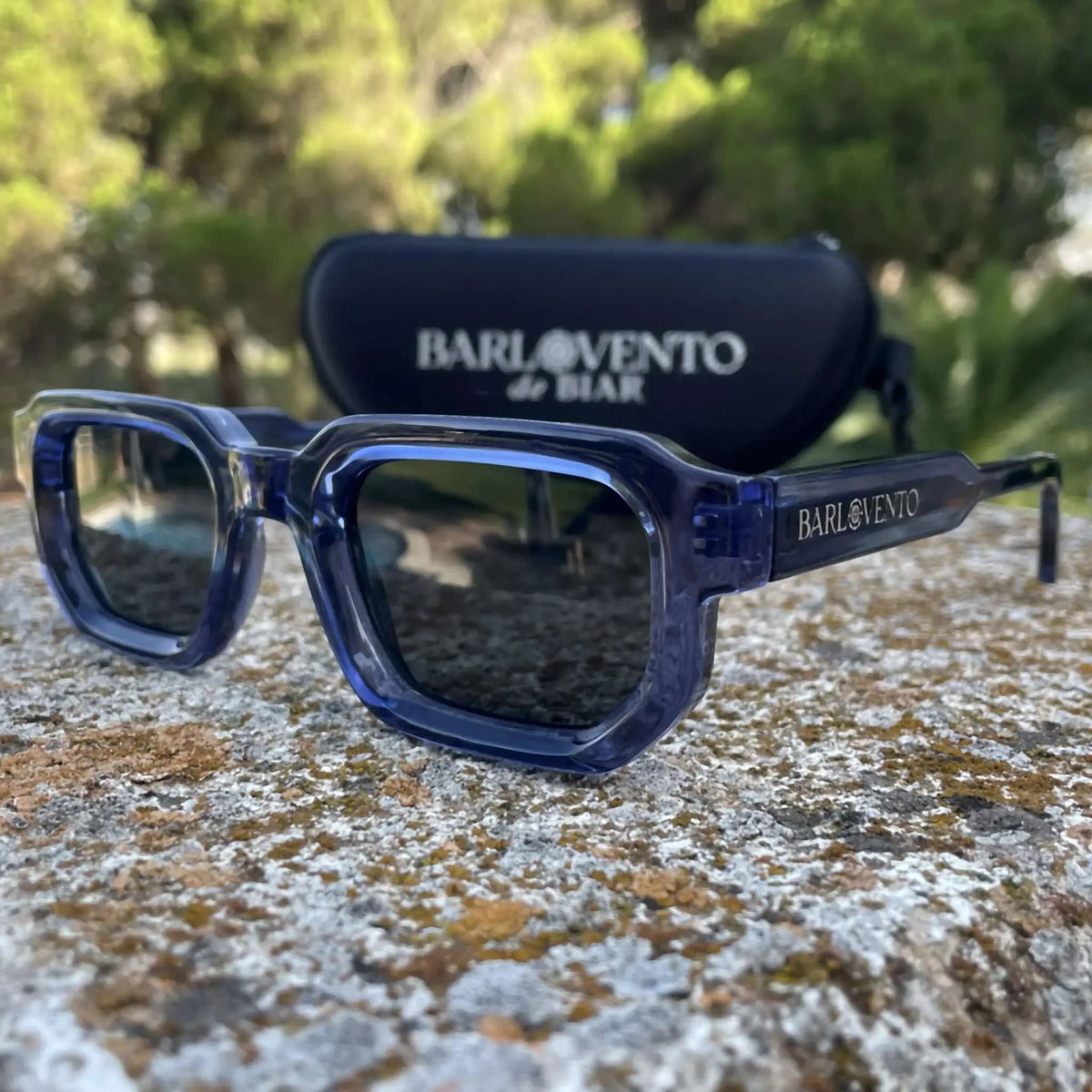 Gafas de sol de montura cuadrada en acetato azul traslúcido con lentes oscuras y estuche negro al fondo.