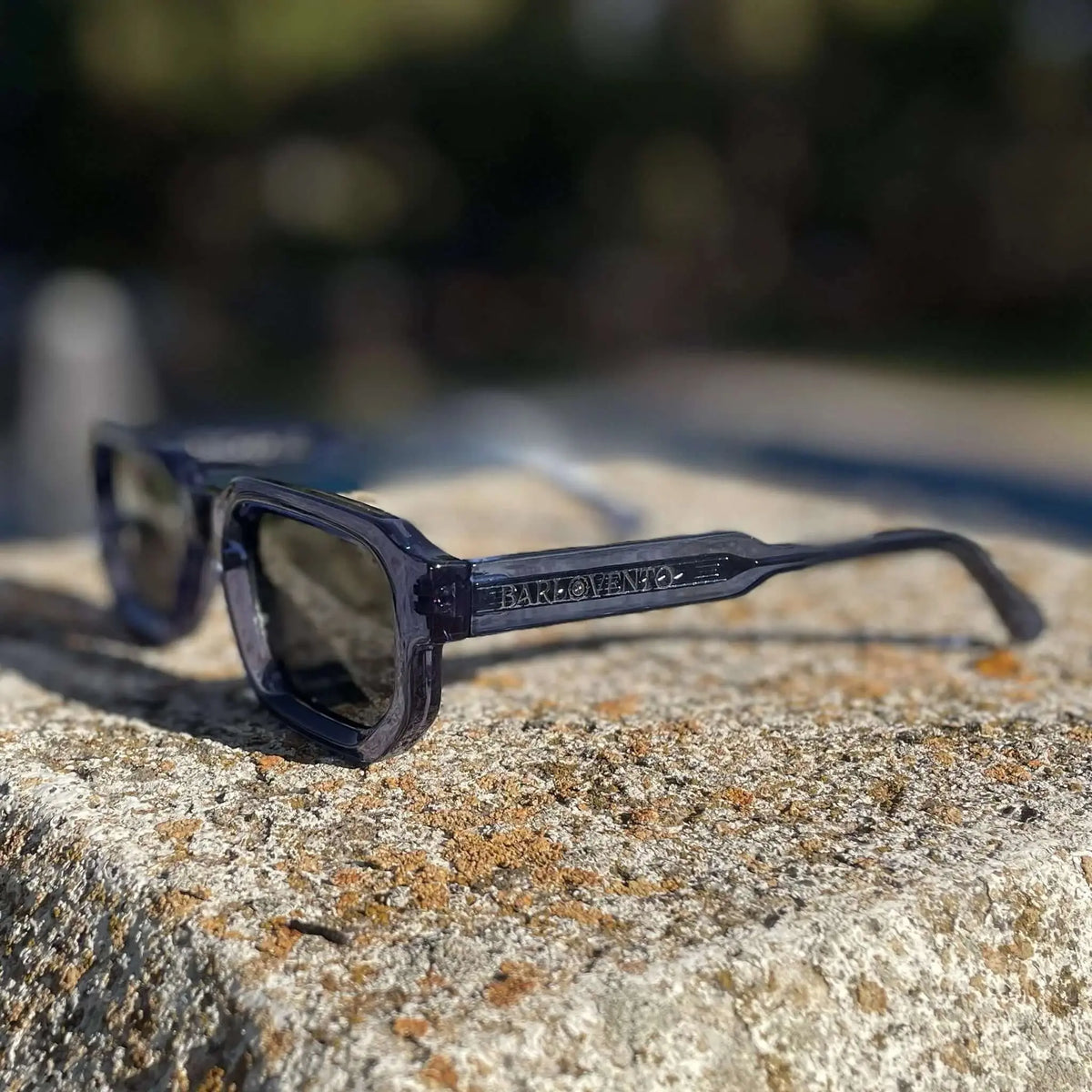 Gafas de sol rectangulares en acetato traslúcido azul, con lentes oscuras, sobre una base de piedra rústica.