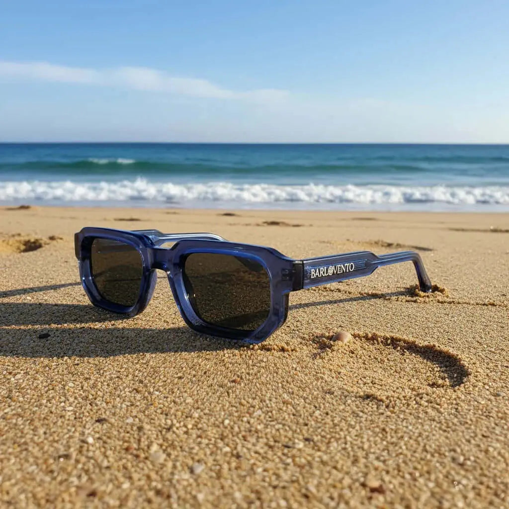 Gafas de sol de montura cuadrada en azul translúcido con lentes oscuras sobre la arena de una playa.
