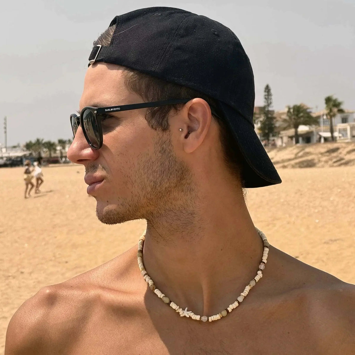 Hombre con gorra negra y gafas de sol de montura negra y lentes verdes polarizadas en la playa. Estilo casual de verano.