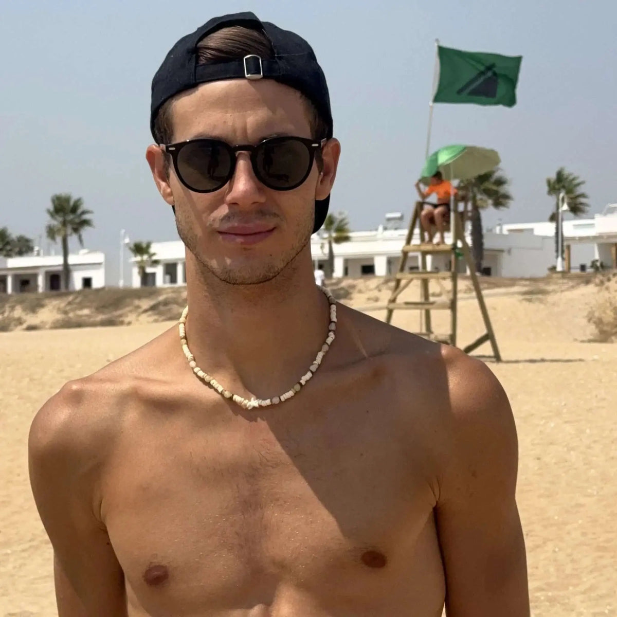 Joven con gorra y gafas de sol negras de montura redonda y lentes verdes polarizadas en una playa soleada.