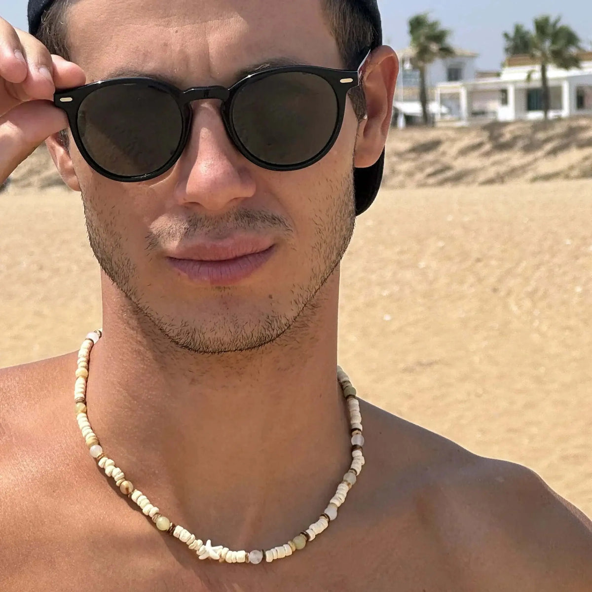 Hombre con gafas de sol negras de montura redonda y lentes verdes polarizadas en la playa con collar de cuentas.