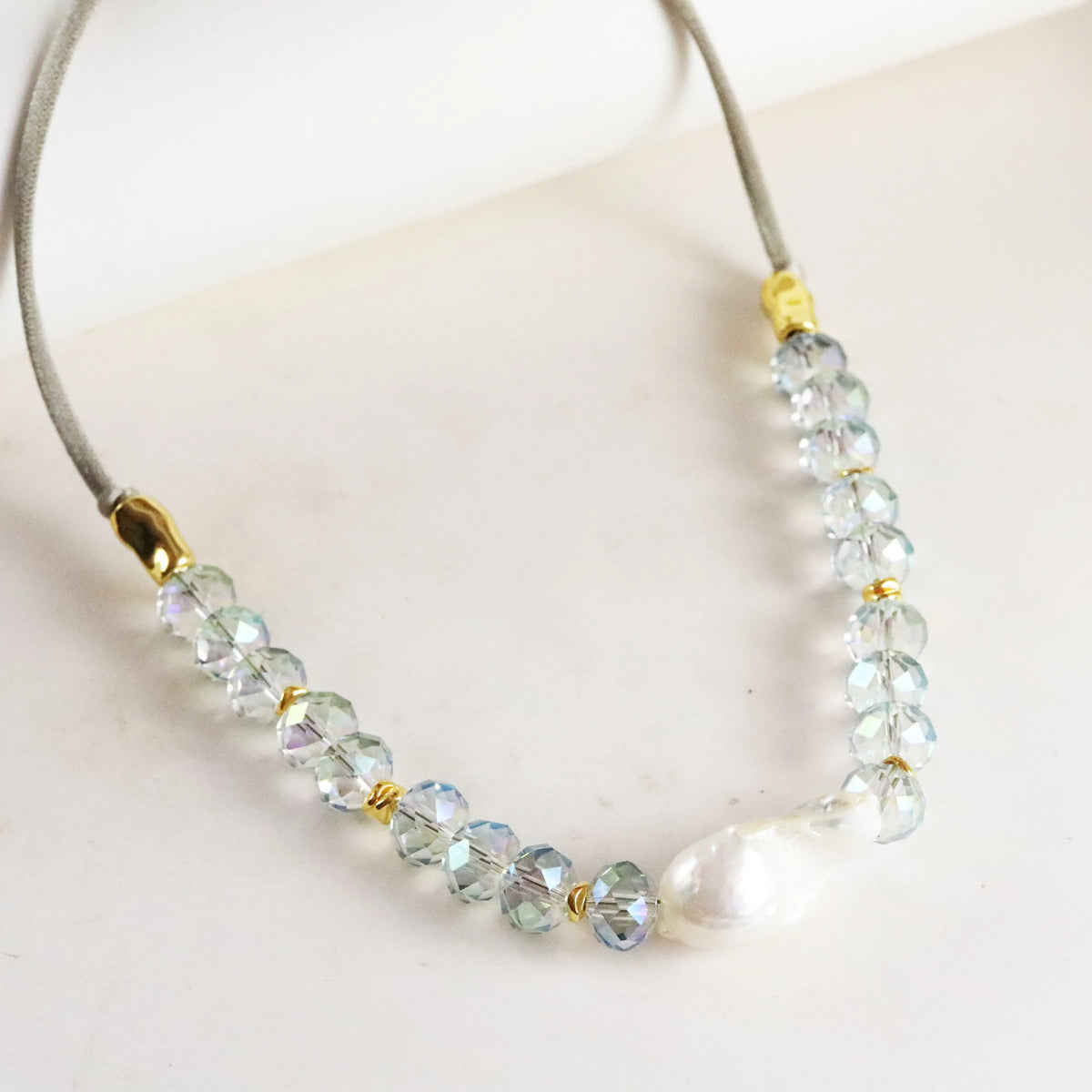 Collar elegante de cristal azul y perla blanca, con detalles en dorado y cordón gris claro.