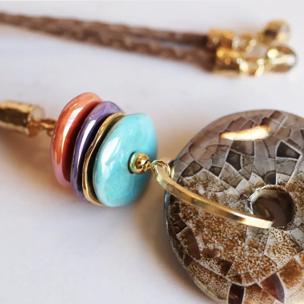 Pendiente bohemio de resina en tonos coral, lila y azul, con detalles dorados y un disco de piedra decorativa.