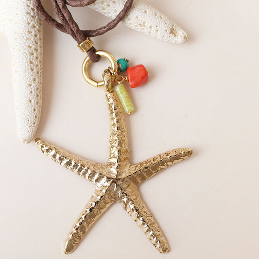 Collar con colgante de estrella de mar dorada, material metálico, con detalles en coral y turquesa sobre cordón marrón.