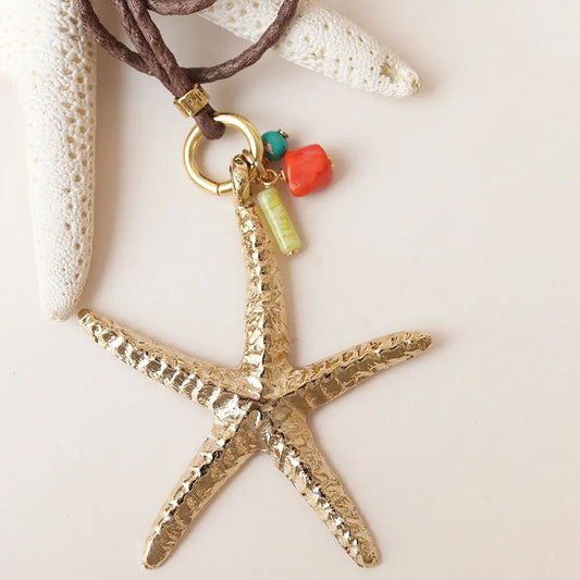 Collar con colgante de estrella de mar dorada, material metálico, con detalles en coral y turquesa sobre cordón marrón.