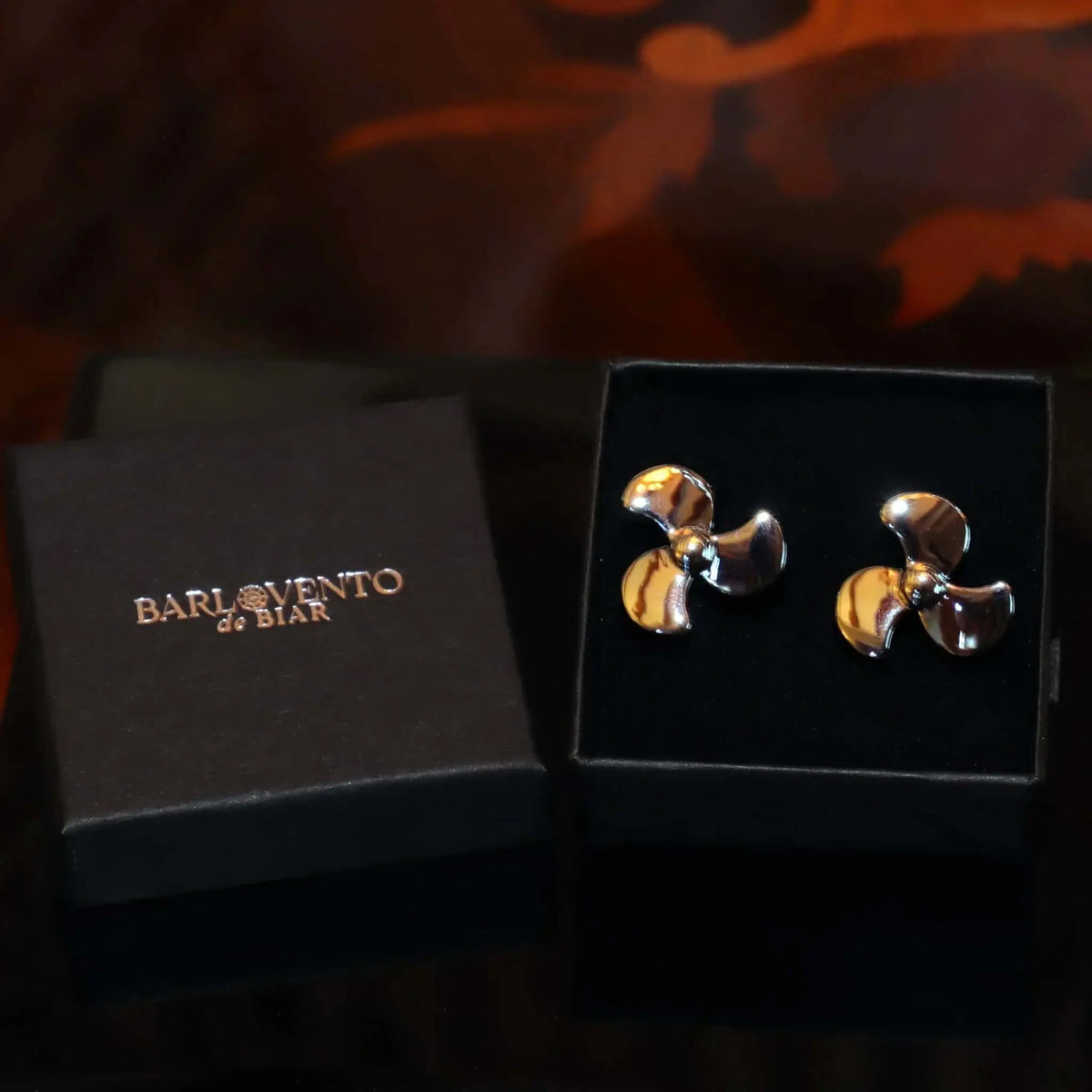 Gemelos para camisa con forma de hélice de barco en metal plateado brillante, presentados en una caja negra.