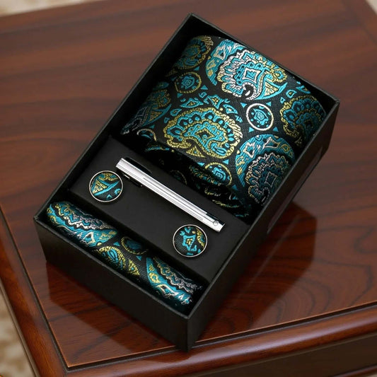 Set de corbata seda paisley verde y azul con pañuelo, gemelos y pasador metálico en caja de regalo sobre mesa madera.
