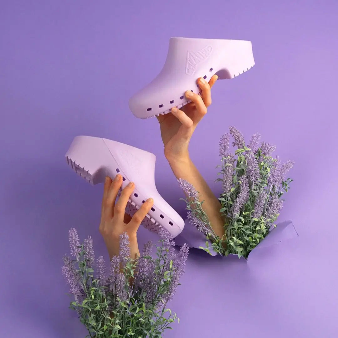 Two hands hold lavender-colored, hole-free slip-on clogs against a purple background with decorative lavender sprigs.