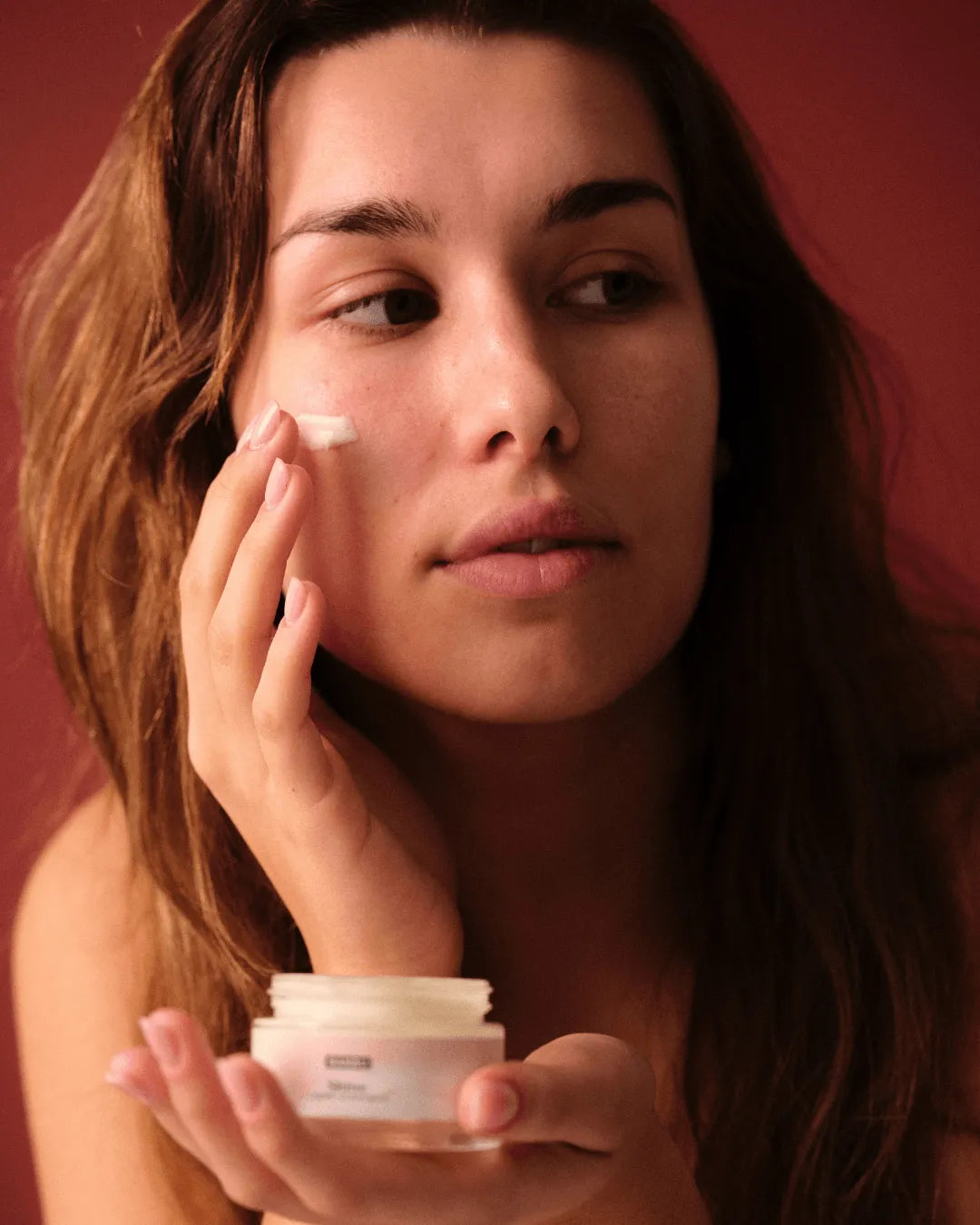 Mujer aplicando la crema facial hidratante Shine de Banbu, de textura suave, frente a un fondo granate cálido.