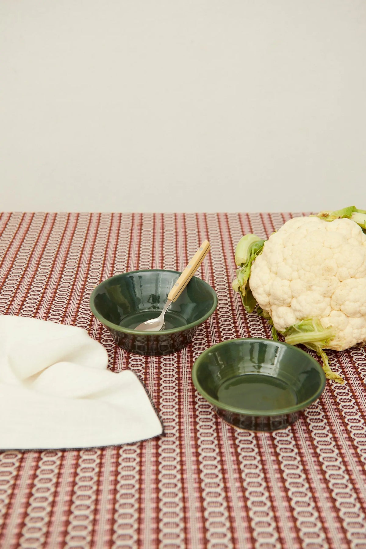 Dos cuencos de cerámica verde brillante sobre mantel estampado, junto a una coliflor y una cuchara.