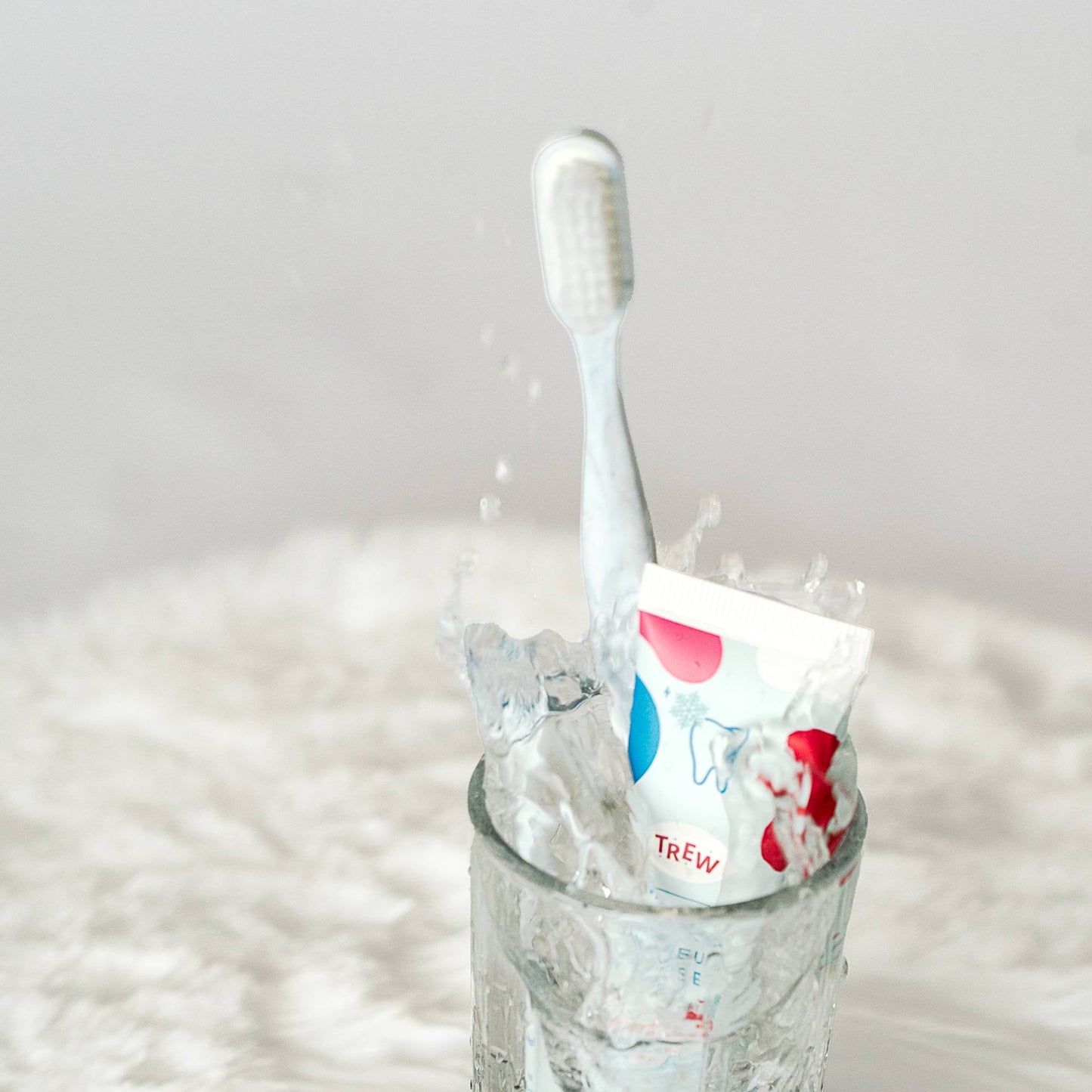 Brosse à dents et tube Trew dans un verre d'eau avec éclaboussures. Design blanc, bleu et rouge, fond texturé blanc.