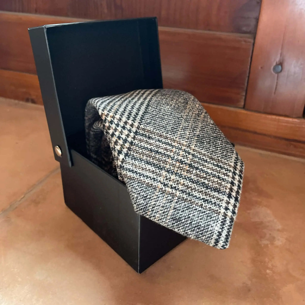 Corbata de lana con patrón de cuadros en tonos marrones y grises dentro de una caja de regalo negra.