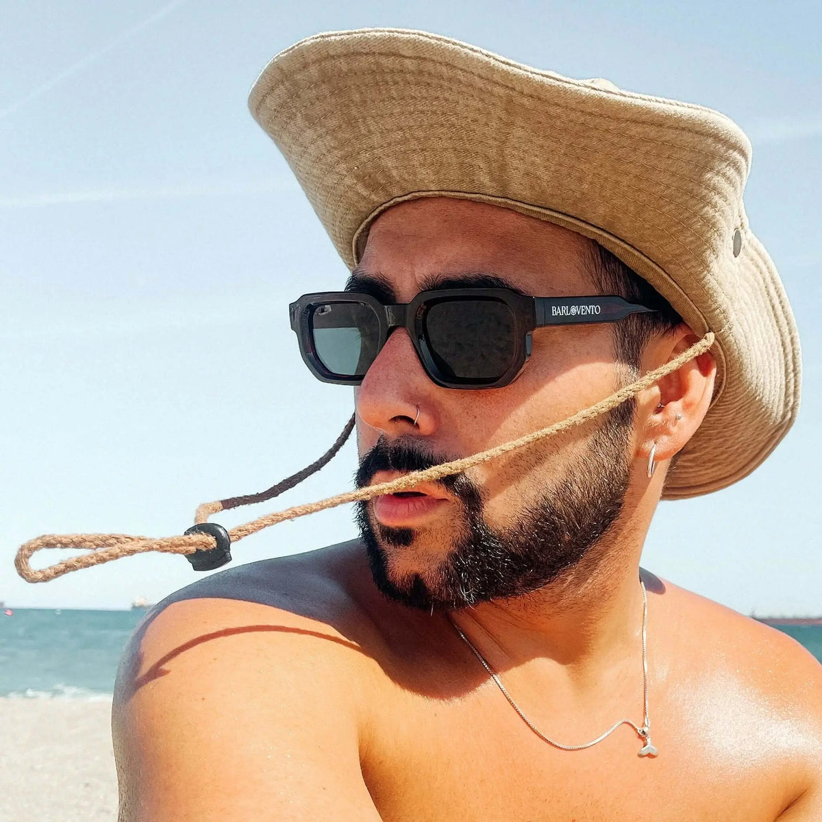 Hombre con sombrero de tela y gafas de sol de montura cuadrada negra y lentes oscuras en la playa.