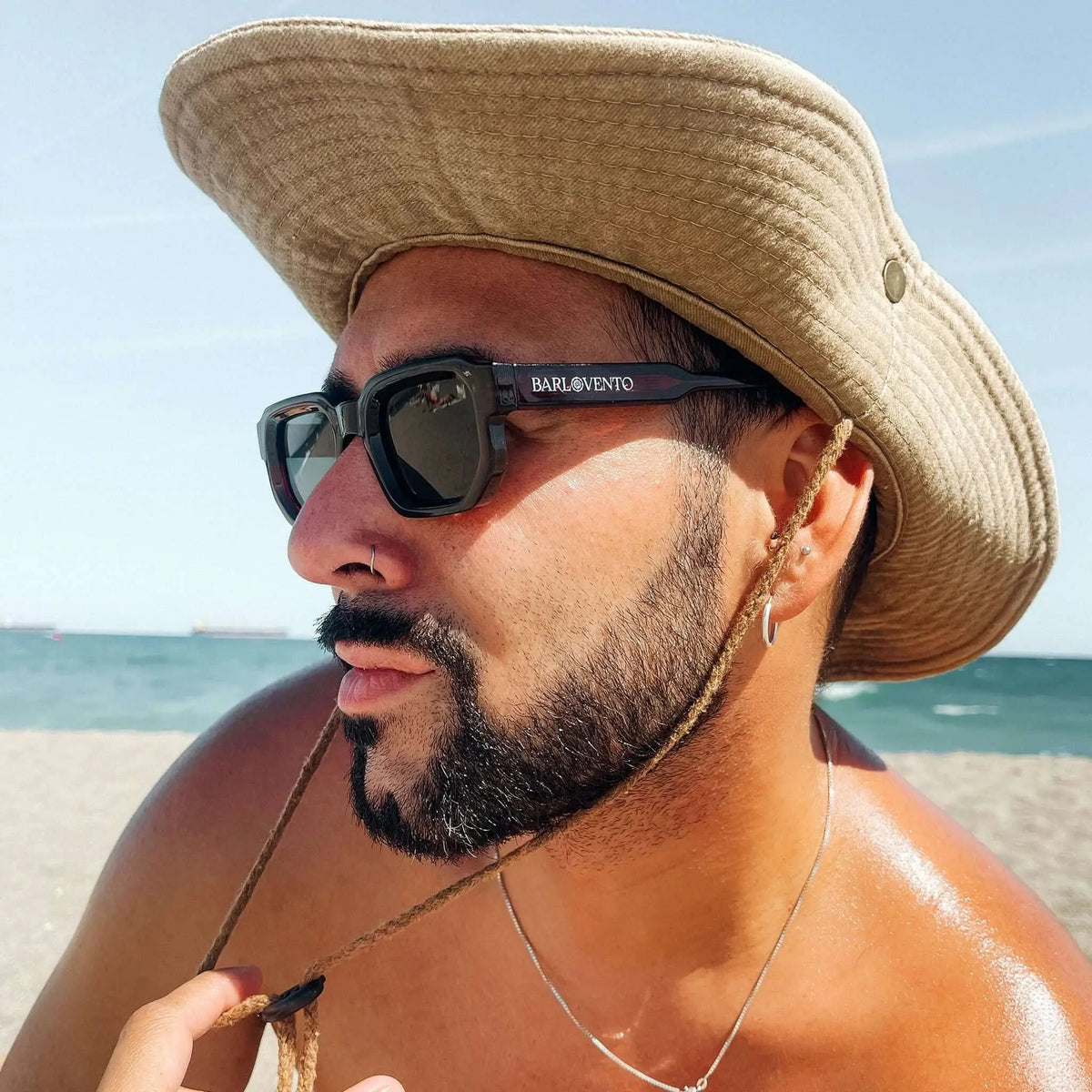 Hombre con sombrero beige y gafas de sol de montura cuadrada color rojo sangre con cristales oscuros en la playa.