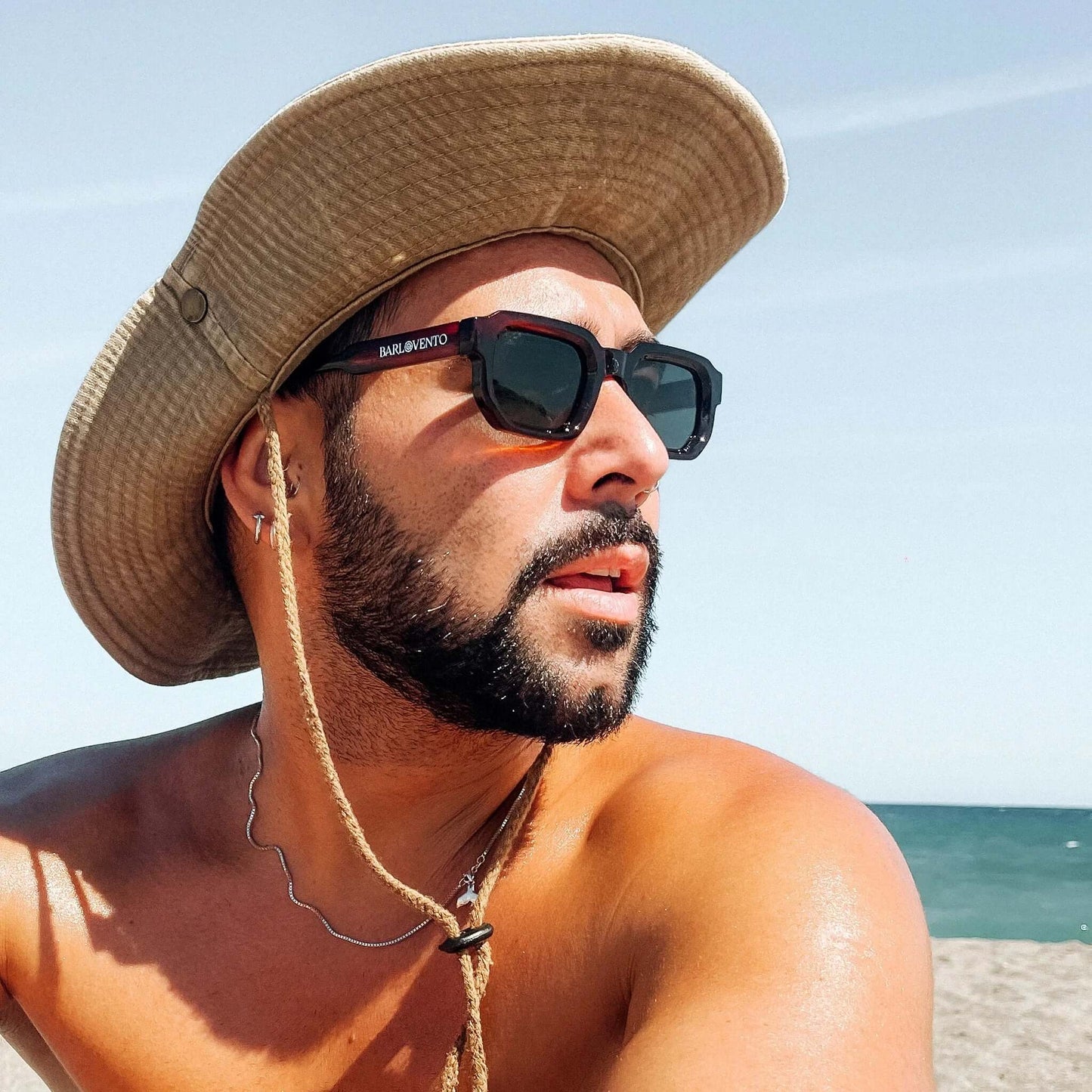 Hombre con sombrero beige y gafas de sol de montura roja oscura cuadrada y cristales oscuros en una playa.