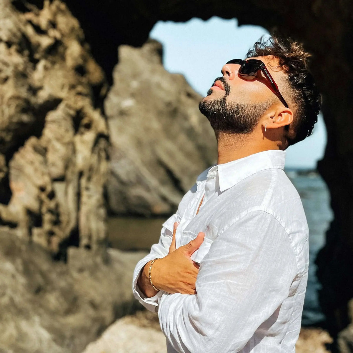 Hombre con camisa blanca usando gafas de sol con montura color rojo sangre y lentes oscuros en un entorno rocoso.