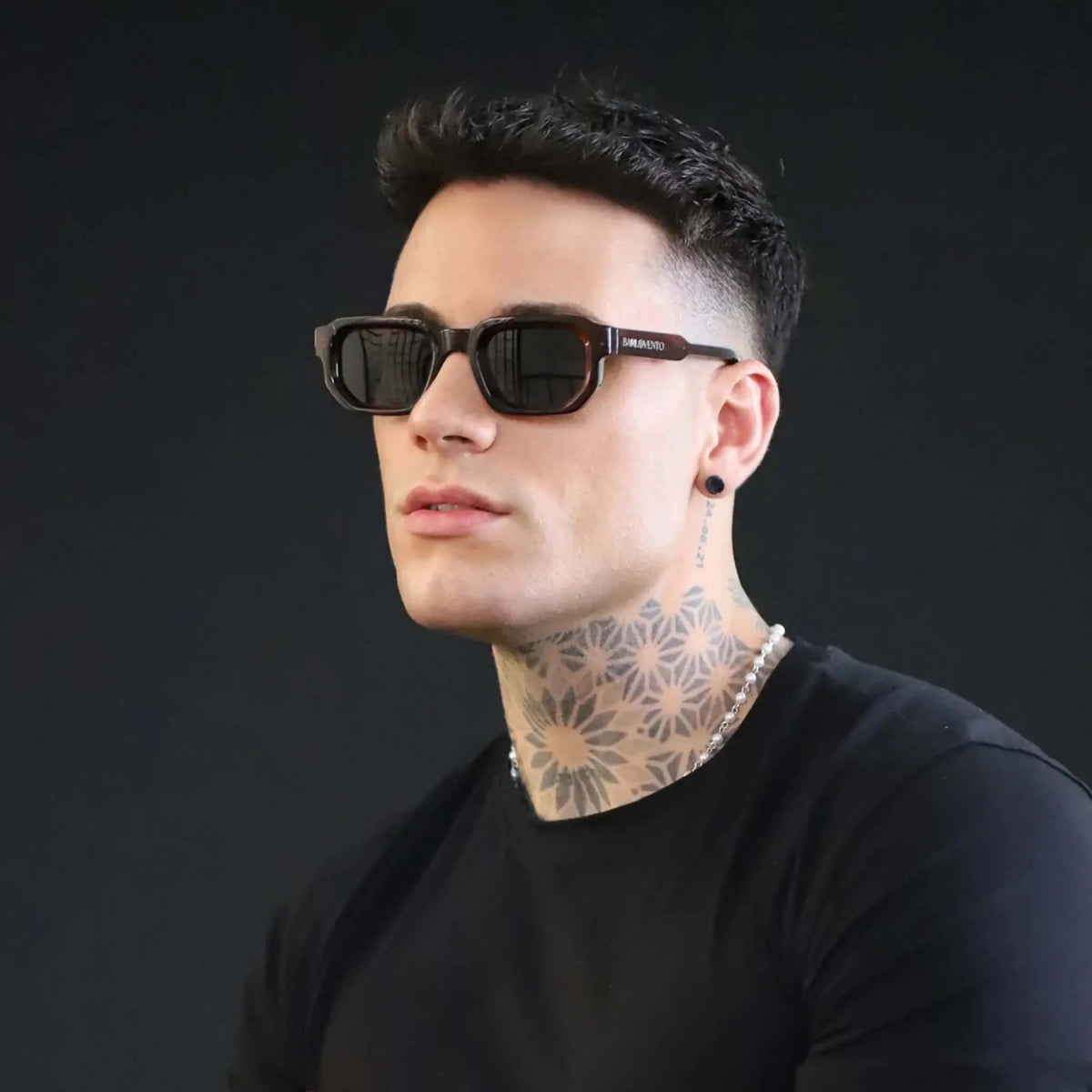 Gafas de sol de montura rectangular estilo carey oscuro con lentes oscuras sobre un hombre con tatuajes en el cuello.