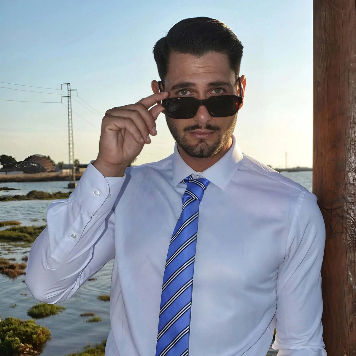 Hombre con camisa blanca, corbata azul y gafas de sol de montura roja oscura y lentes oscuras al aire libre.
