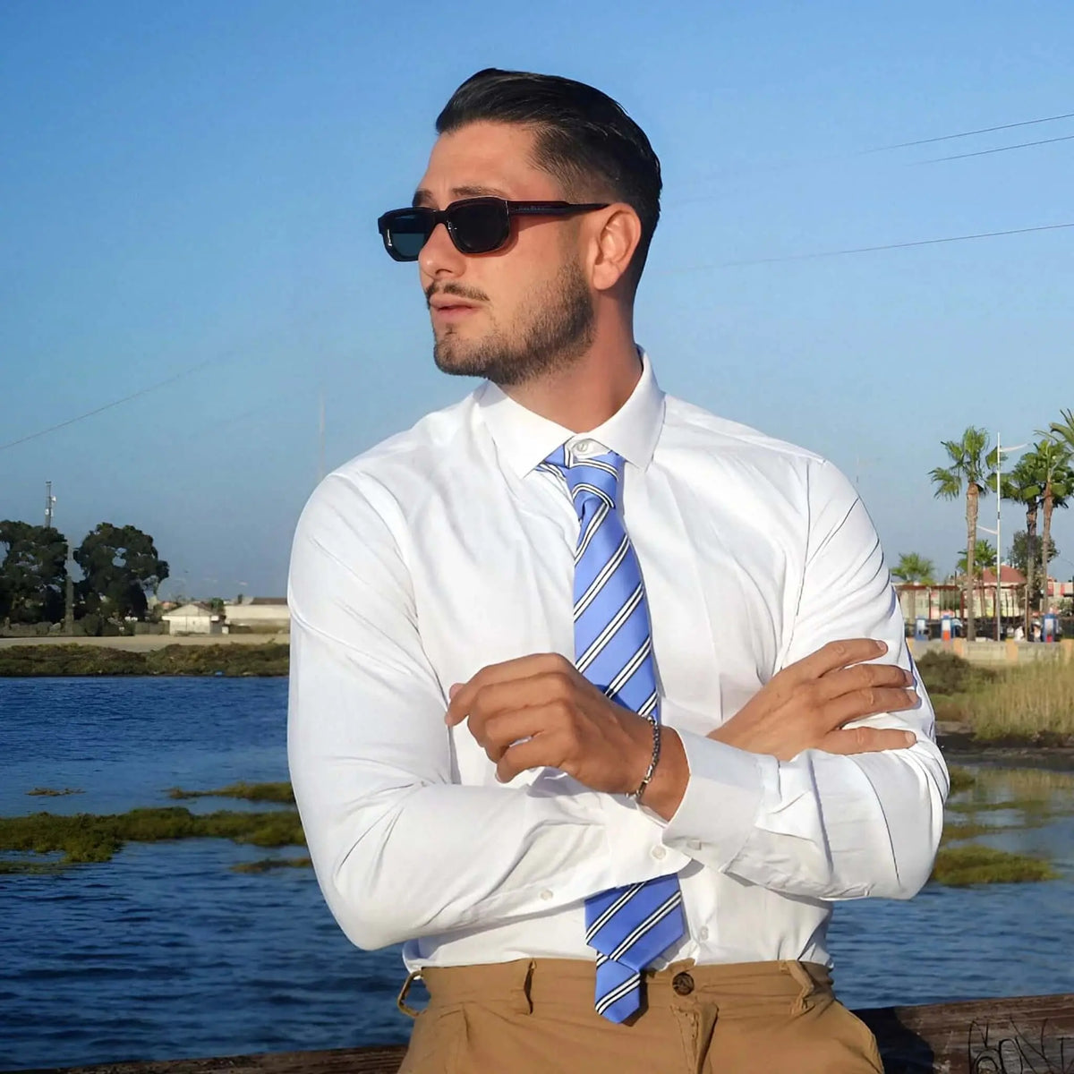 Hombre con camisa blanca y corbata azul luce gafas de sol de montura roja y lentes oscuros en un paisaje costero.