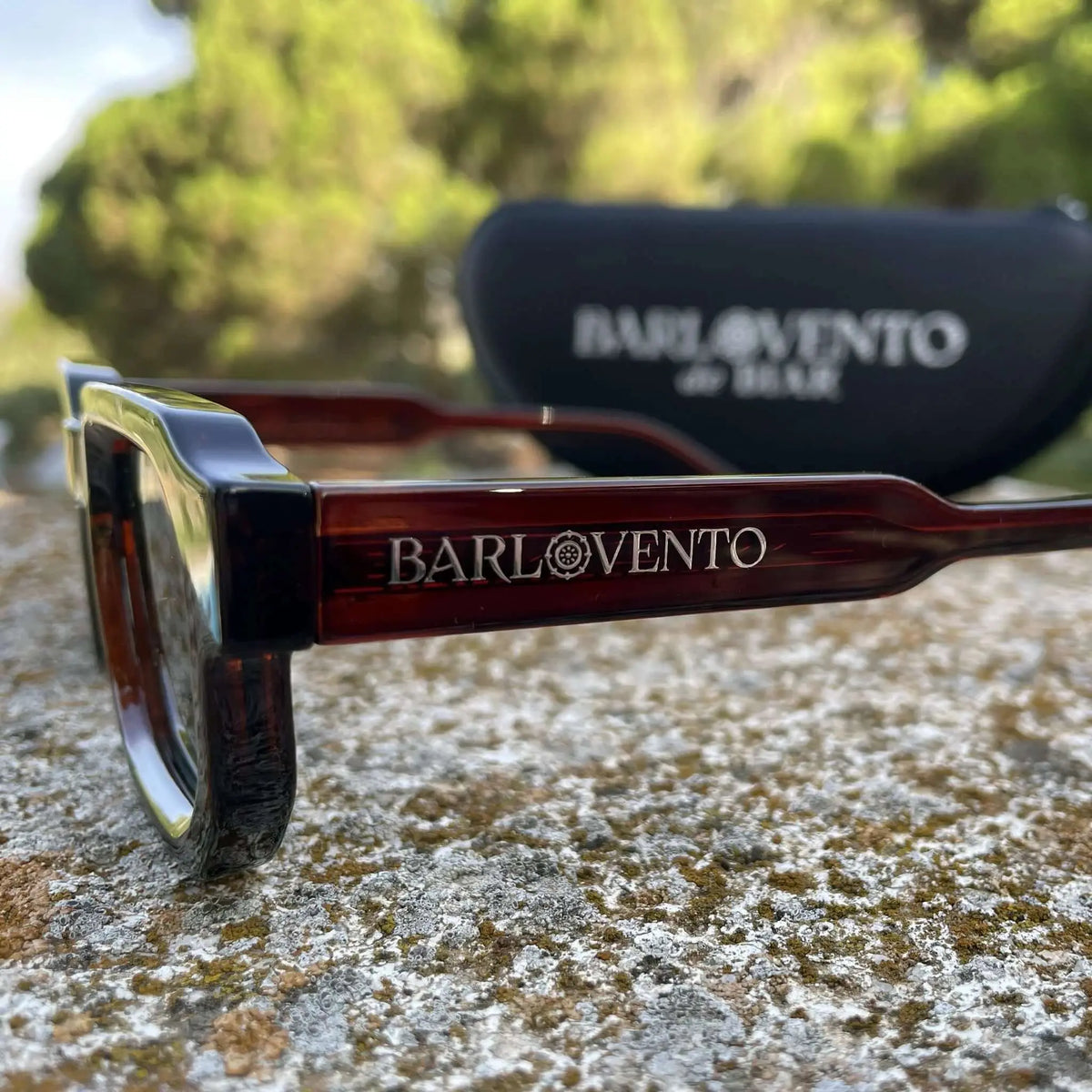 Gafas de sol de pasta color rojo oscuro traslúcido y lentes verdes, estilo rectangular, sobre una superficie de piedra.