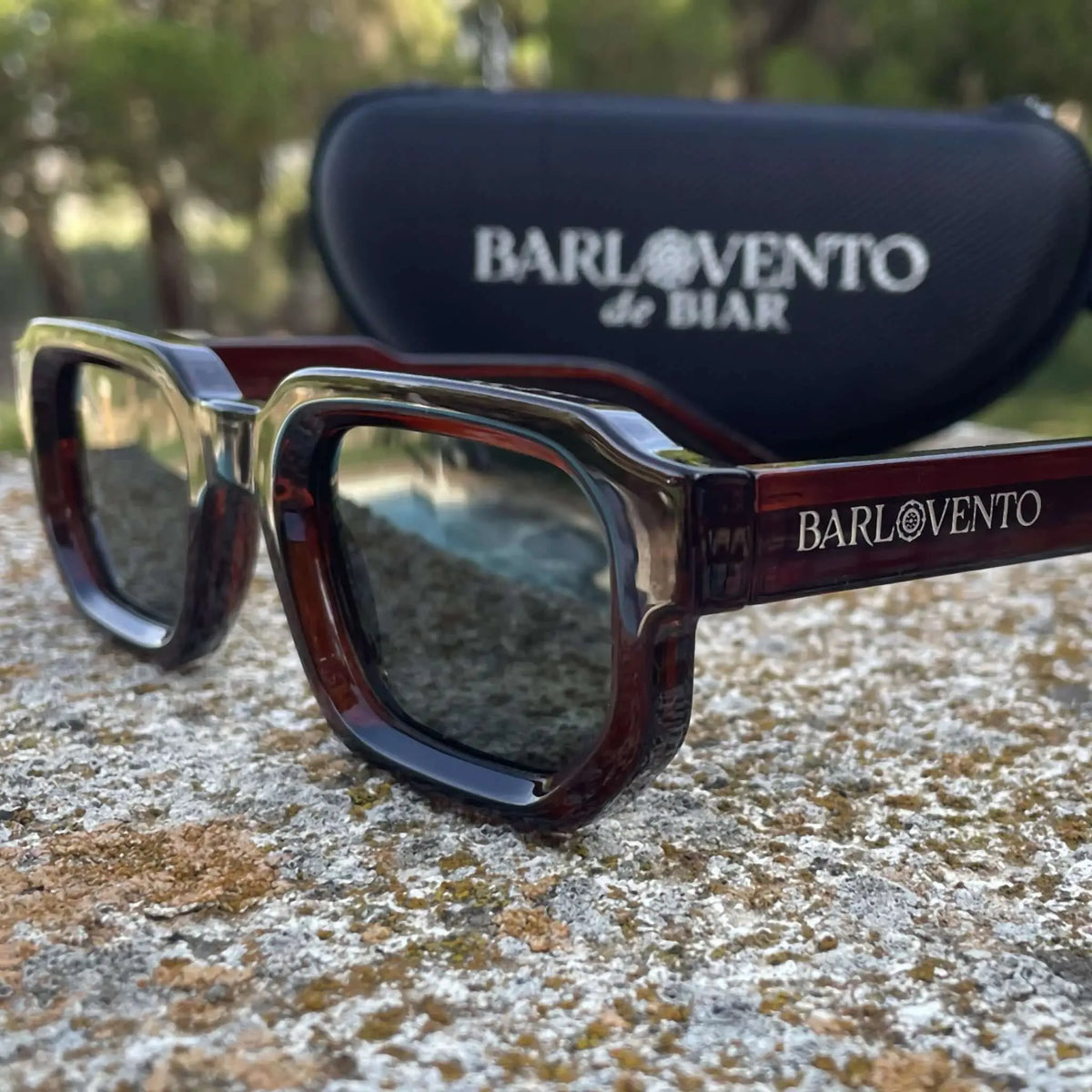 Gafas de sol rectangulares color rojo sangre con lentes oscuros, sobre una superficie de piedra y estuche de fondo.