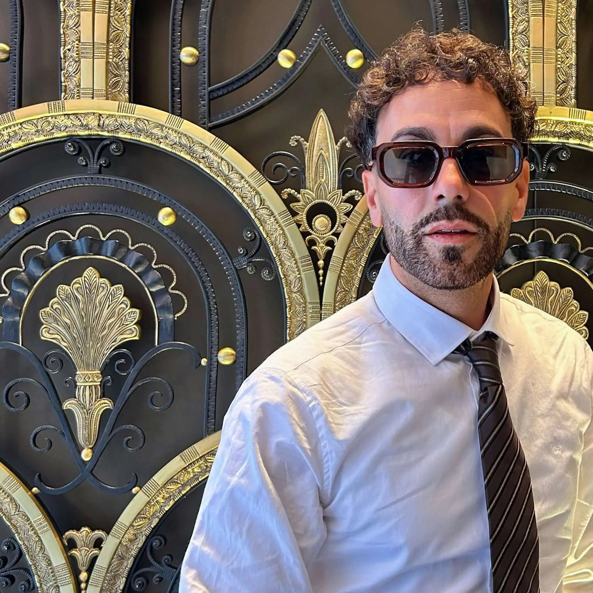 Hombre con gafas de sol de montura rectangular rojiza y lentes oscuras, camisa blanca y corbata ante una puerta ornate.