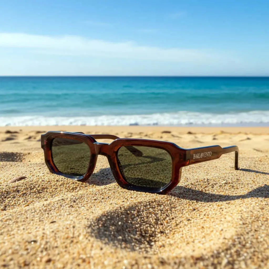 Gafas de sol rectangulares con montura de acetato color café translúcido y lentes oscuros, sobre la arena de una playa.