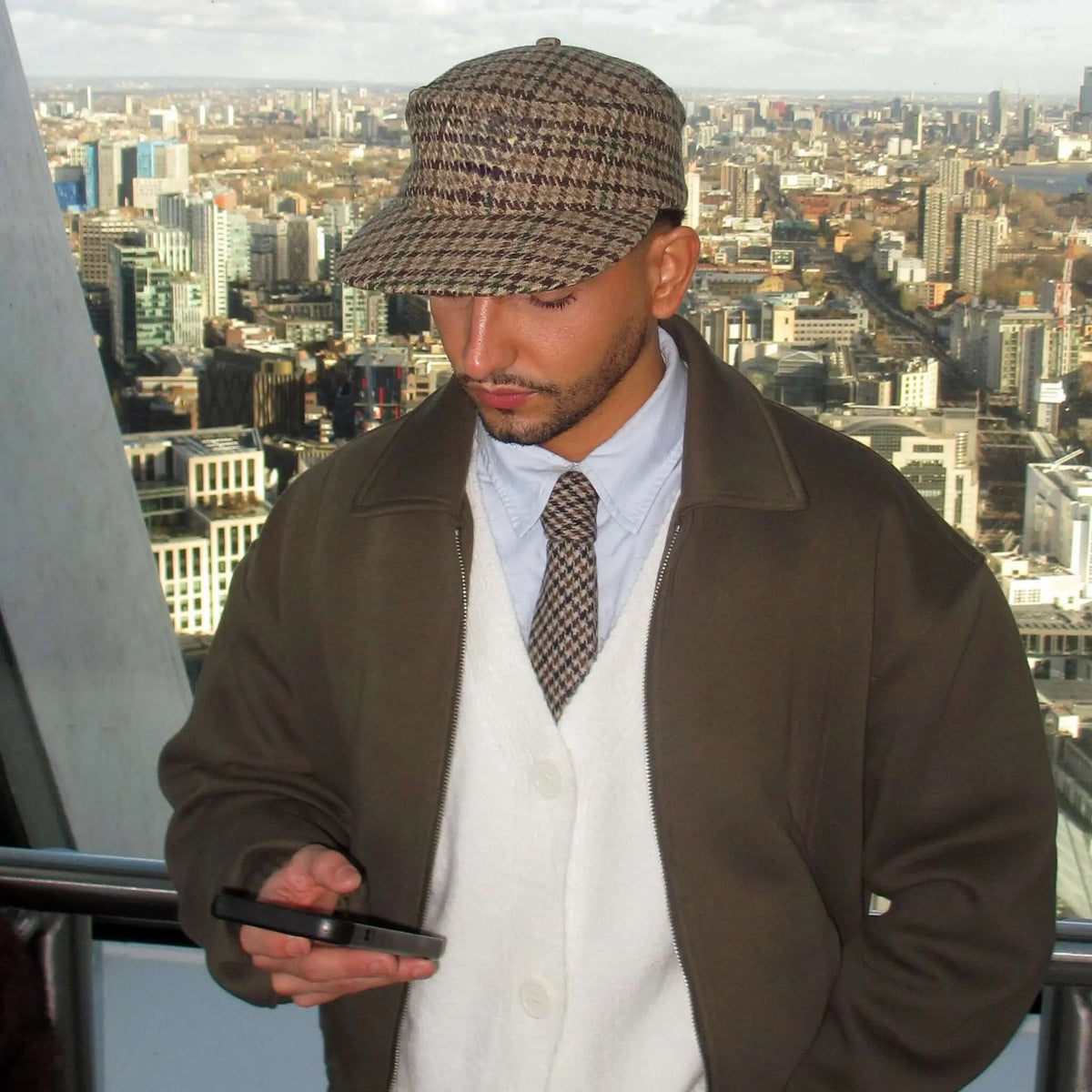 Hombre con corbata de lana con patrón de pata de gallo, chaqueta marrón, cárdigan blanco y gorra de cuadros.