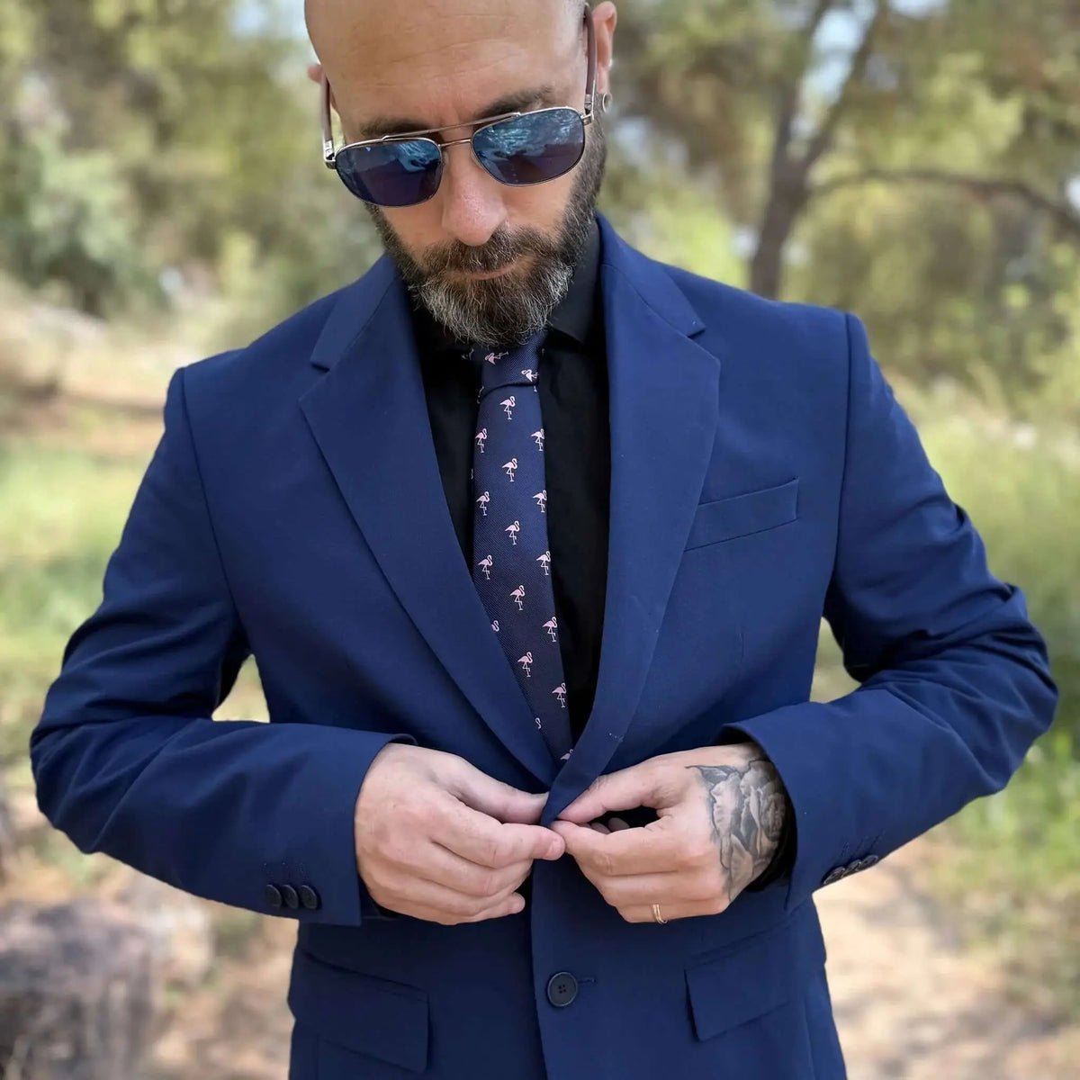Hombre con traje azul, camisa negra y corbata delgada azul marino con estampado de flamencos rosas en exterior.