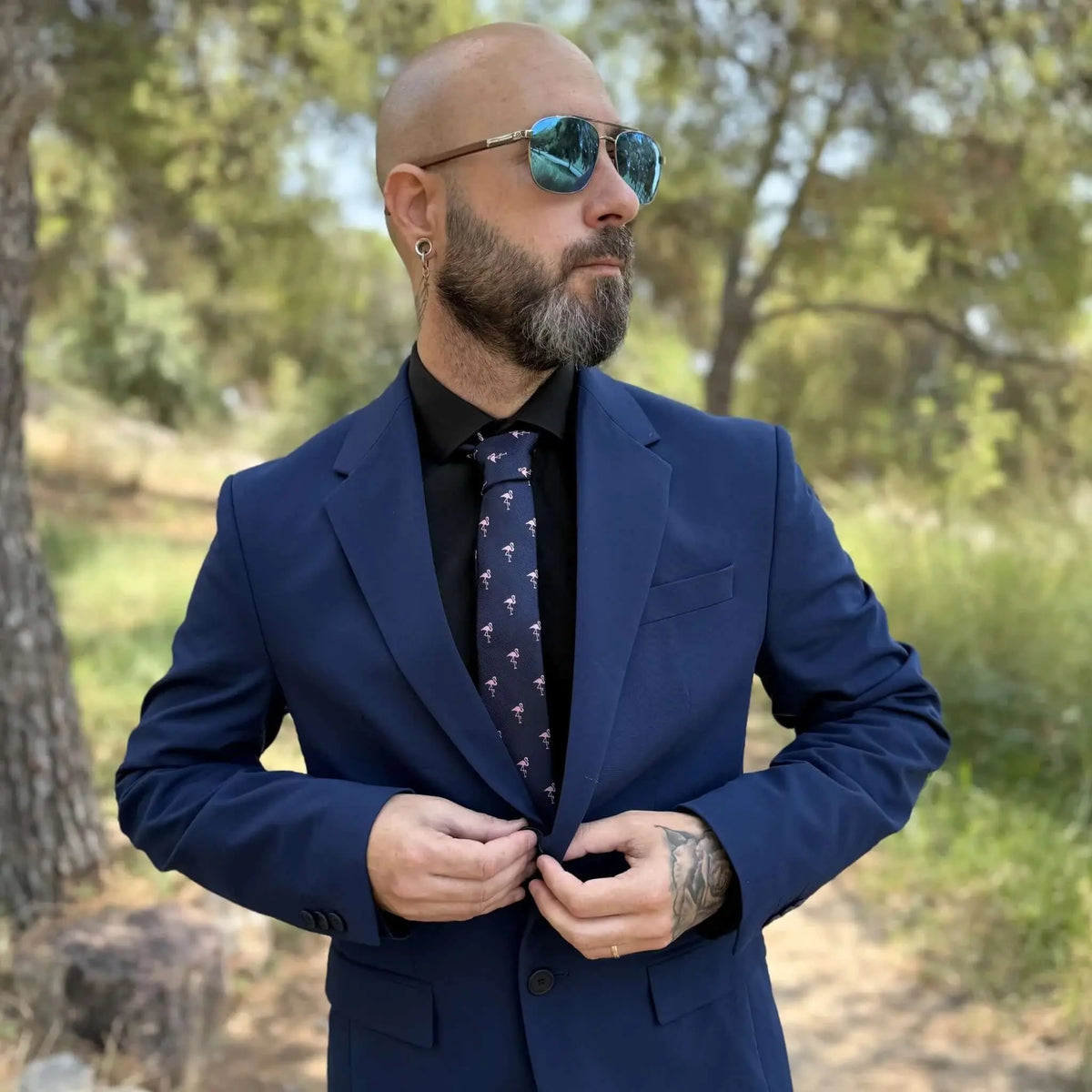 Hombre con traje azul, camisa negra y corbata delgada azul marino con estampado de flamencos rosas en entorno natural.