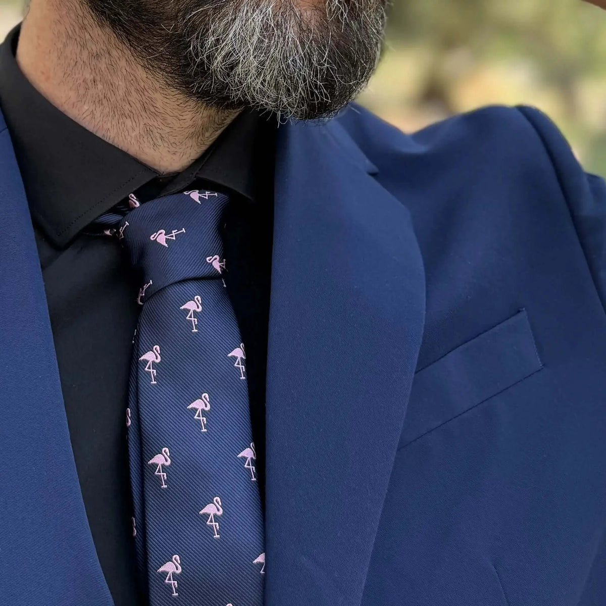Corbata delgada azul marino con estampado de flamencos rosas, sobre camisa negra y traje azul. Estilo formal moderno.