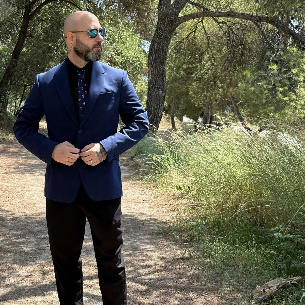 Hombre con americana azul, camisa negra y corbata fina estampada en un entorno natural boscoso.