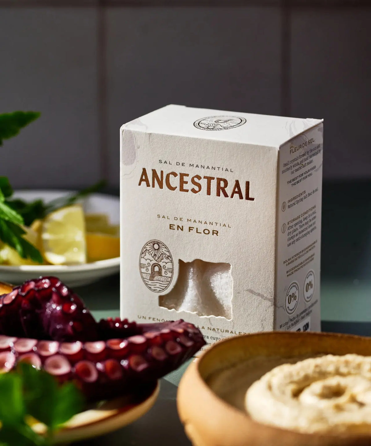 A box of Ancestral spring salt with a cutout window showing salt crystals, placed near octopus and a ceramic bowl.
