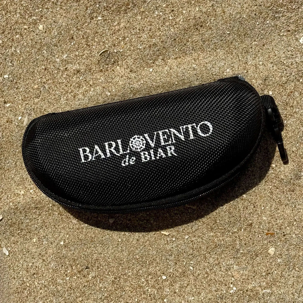 Estuche rígido negro de tela texturizada para gafas de sol, forma ovalada con cremallera, apoyado sobre arena de playa.