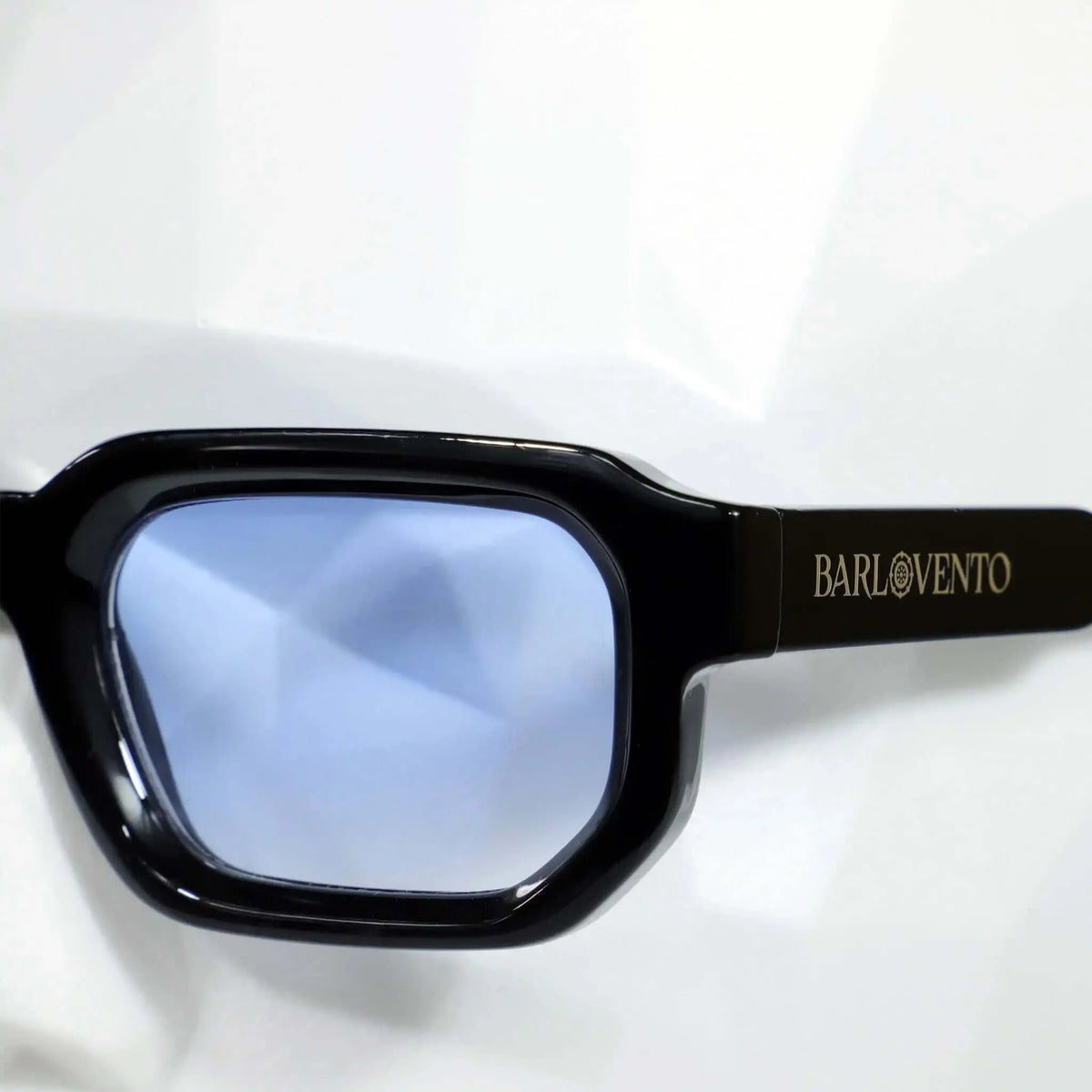 Gafas de sol de montura negra rectangular gruesa con lentes de color azul claro sobre fondo blanco.