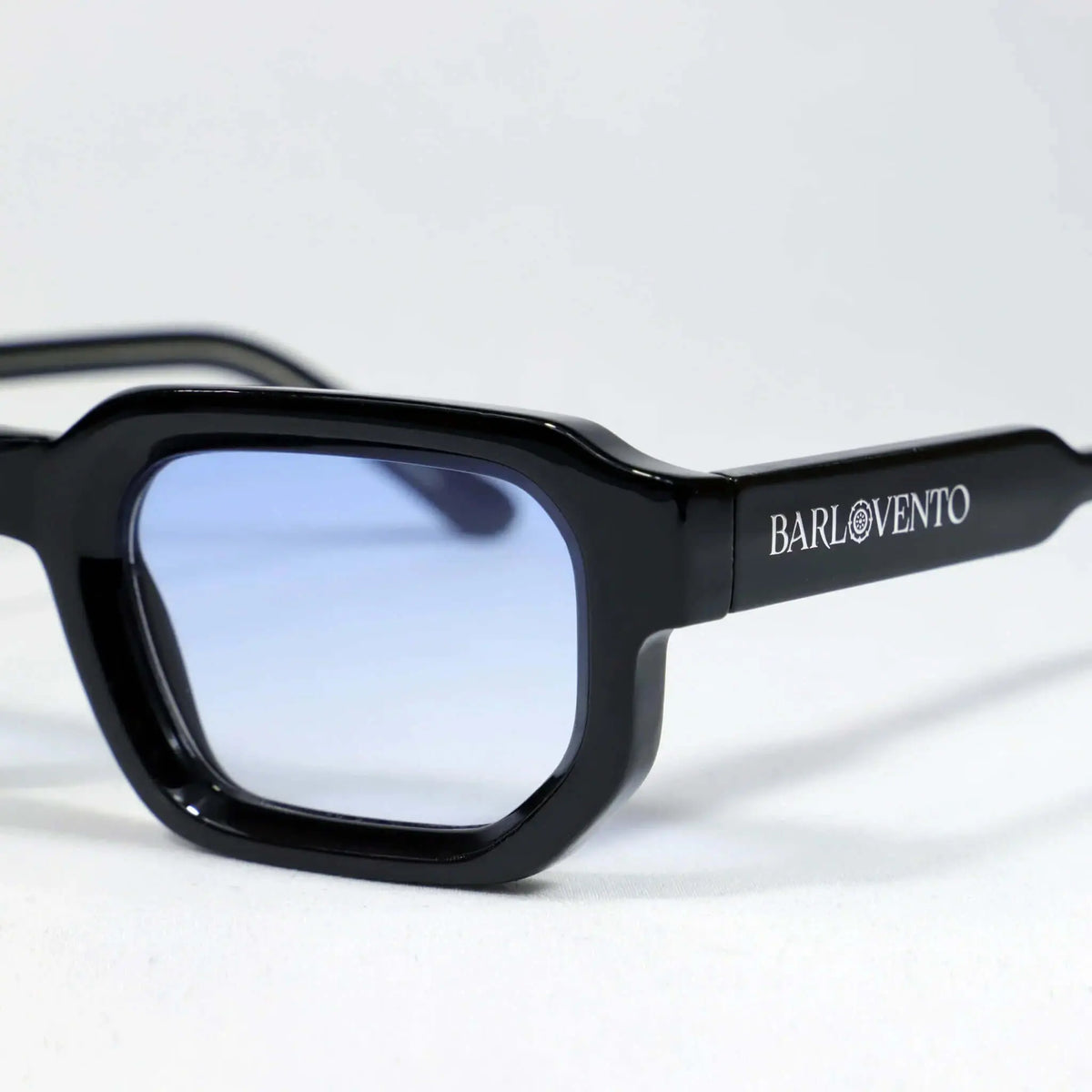Gafas de sol de montura rectangular negra brillante con cristales tintados en azul claro. Diseño moderno y minimalista.
