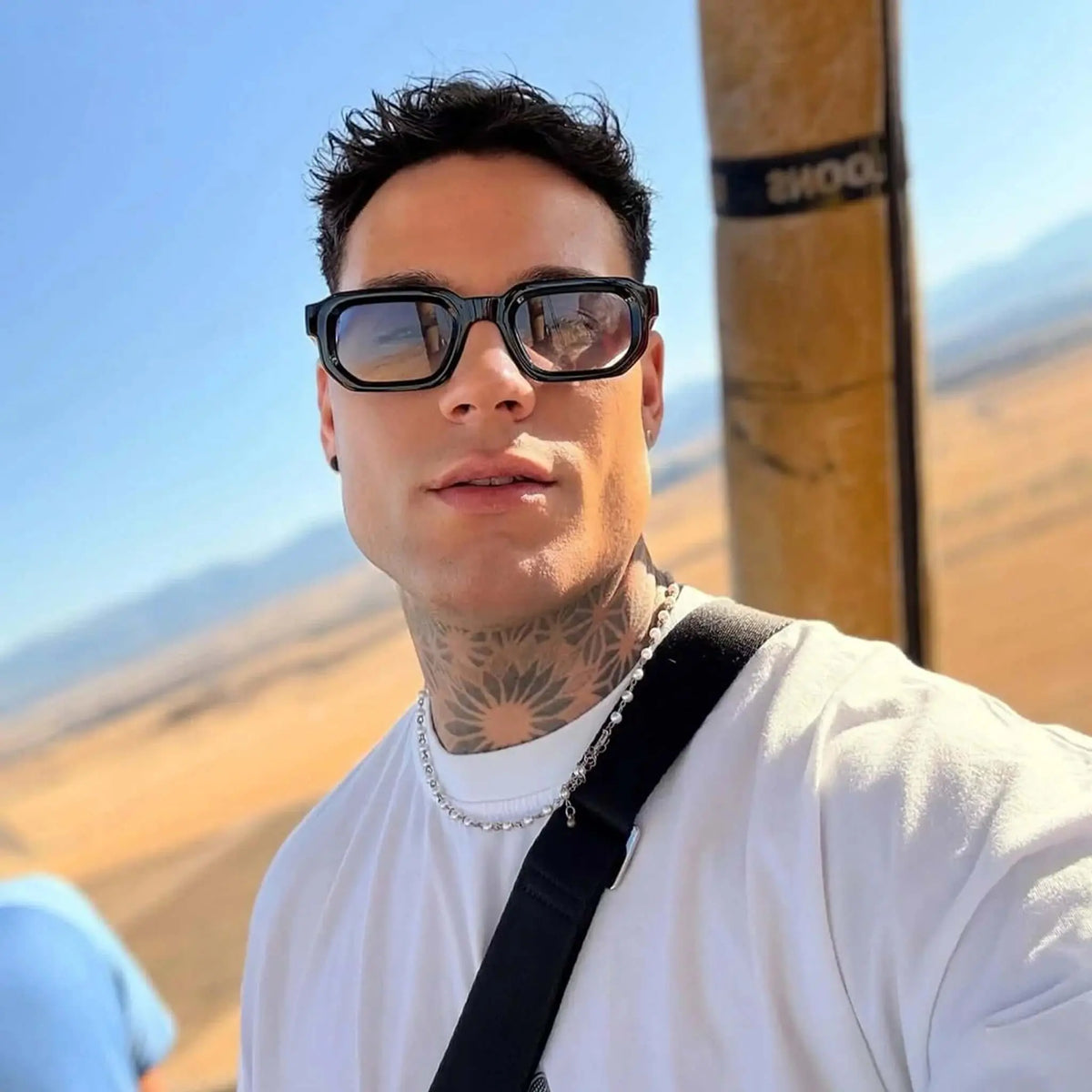 Hombre con gafas de sol de montura negra rectangular y lentes azulados, estilo moderno en un paisaje soleado.