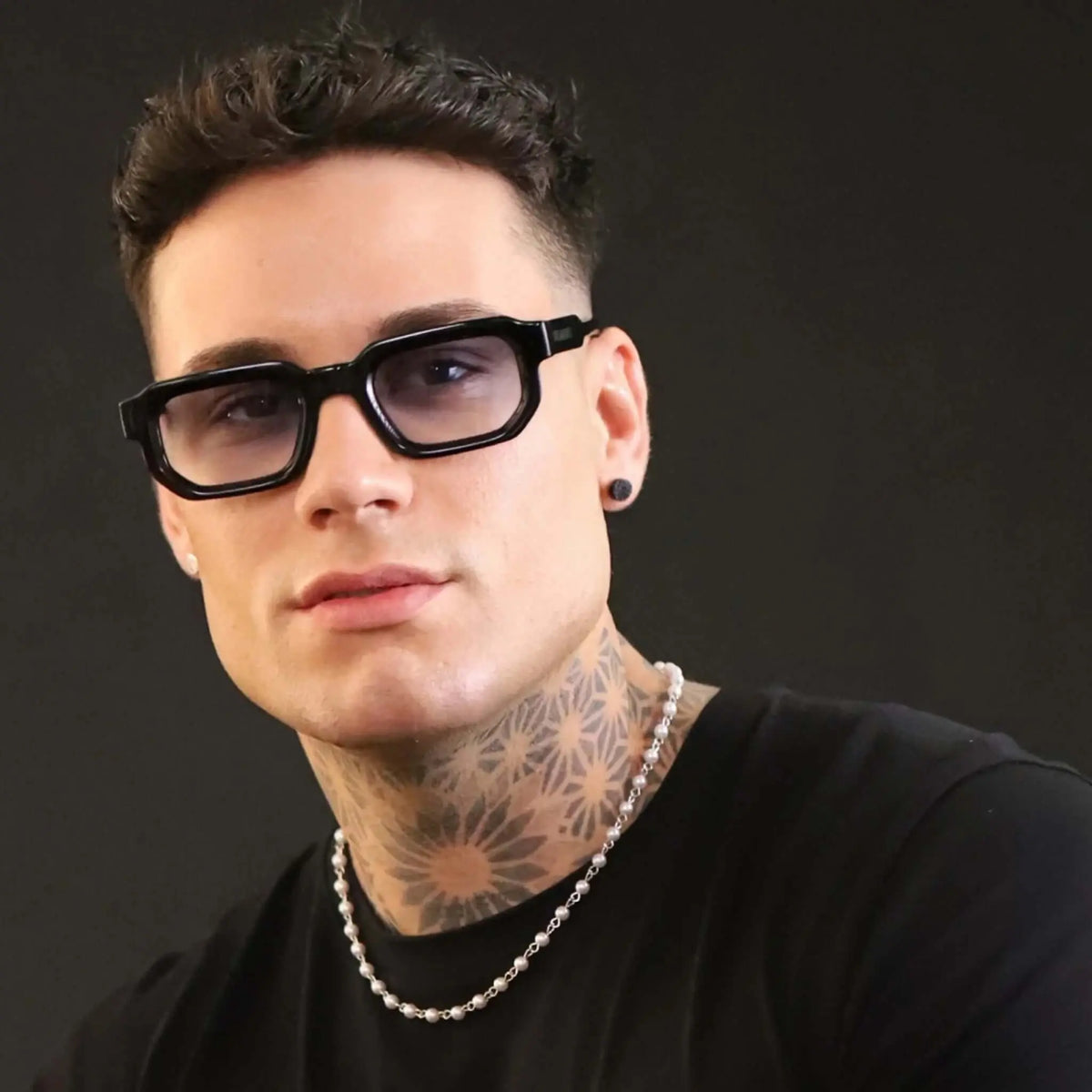 Gafas de sol rectangulares con montura negra gruesa y lentes degradadas en tono azul sobre hombre con tatuajes.